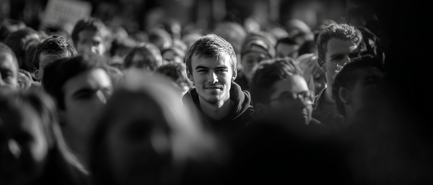 A black-and-white wide shot of a male rally crowd, faces partially visible. The political performance that goes with administrative actions but is not where the actions actually happen.